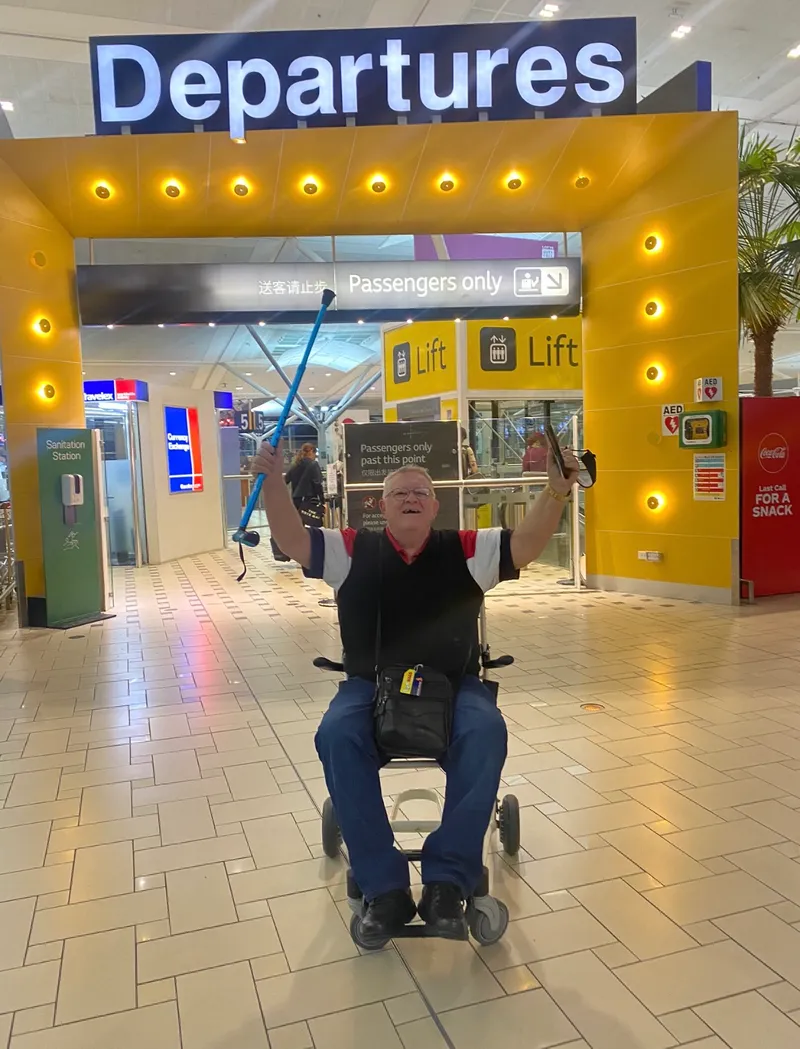 A participant in his wheelchair under the Departures sign at Brisbane Airport, both arms in the air, walking stick held high