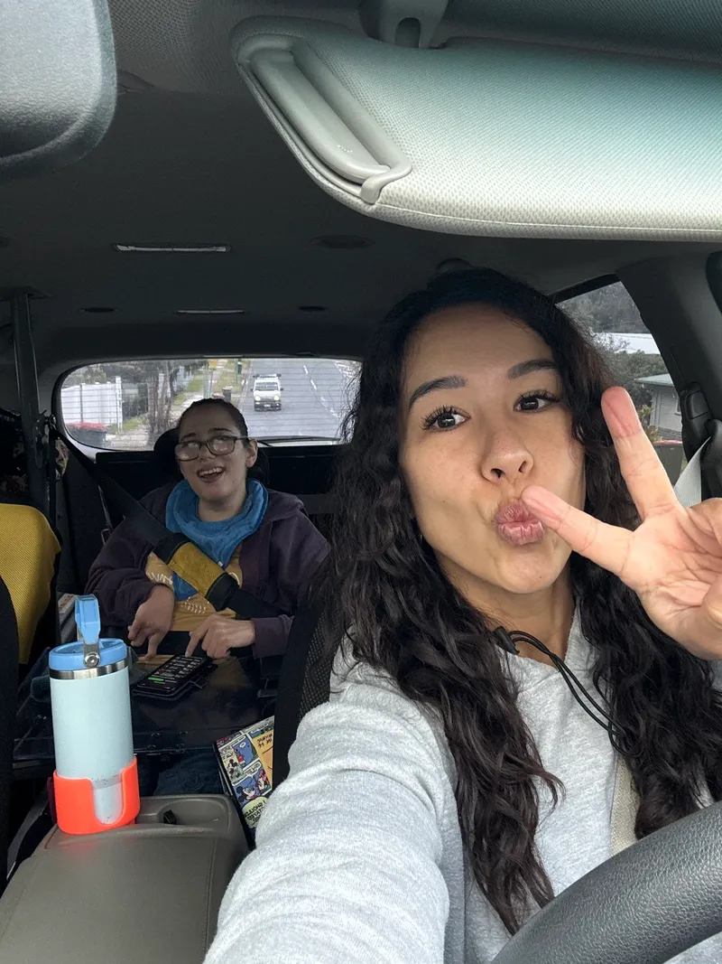 Selfie from the driver's seat: a support worker pulling a face and a peace sign, her participant in the back seat with a tablet, both belted in and parked
