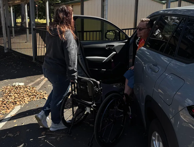 A support worker folding down a wheelchair beside an open car door while a participant settles into the seat, in a sunny car park
