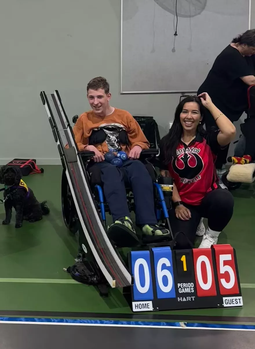 A young man in his wheelchair with his assistance dog beside him, Heidi crouched next to them on the boccia court