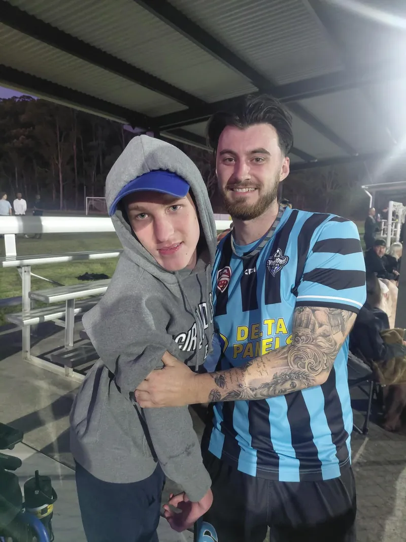 A participant in a grey hoodie and blue cap, arm in arm with a footballer in a blue and black striped kit, at an evening game