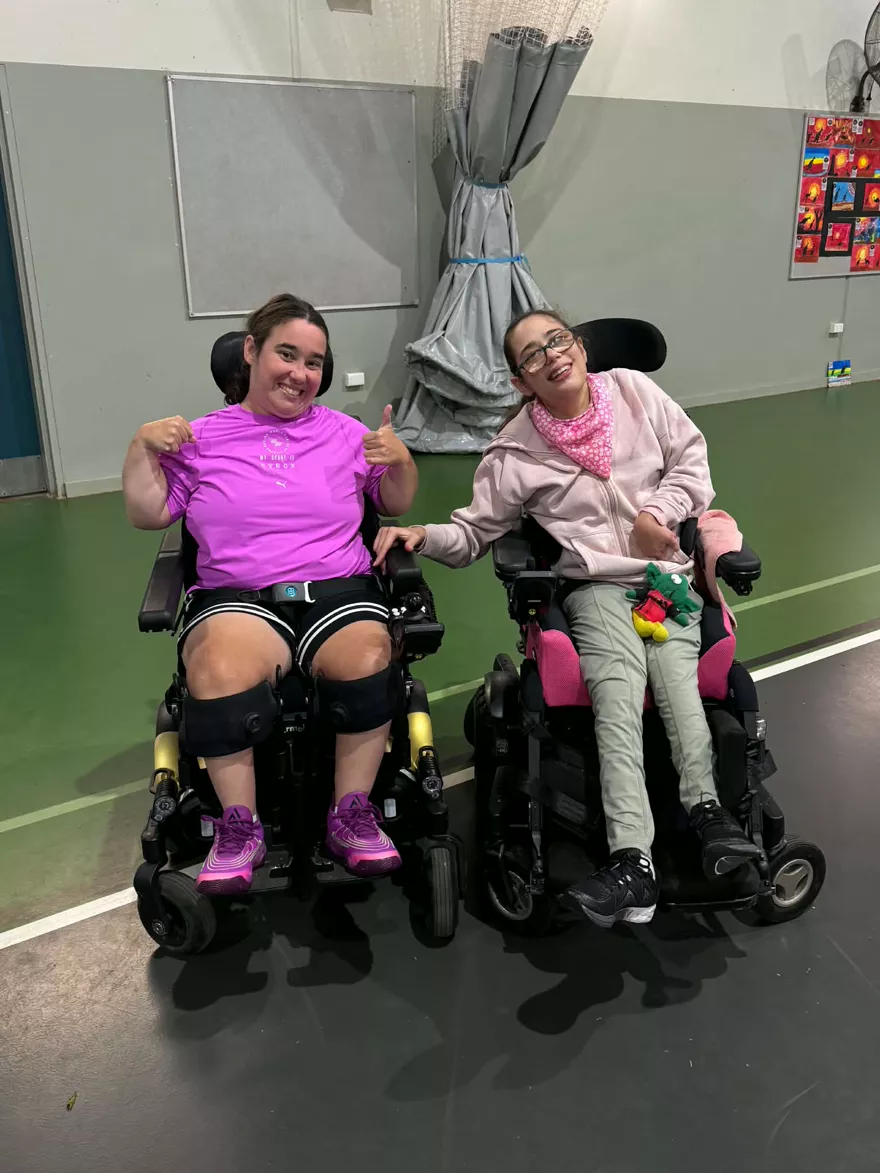 Two participants in their wheelchairs at the sports hall, one giving a thumbs up
