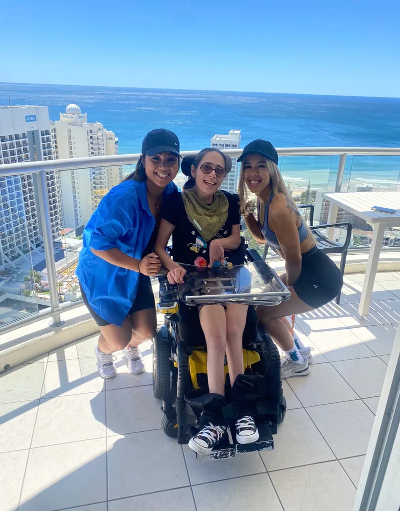 A participant in her powered wheelchair with two support workers either side of her on a sunny balcony, the ocean behind them