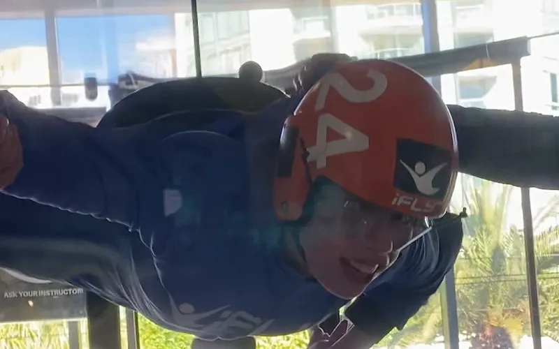 A participant in an orange helmet and blue flight suit flying in the iFLY wind tunnel at Surfers Paradise, arms out, grinning