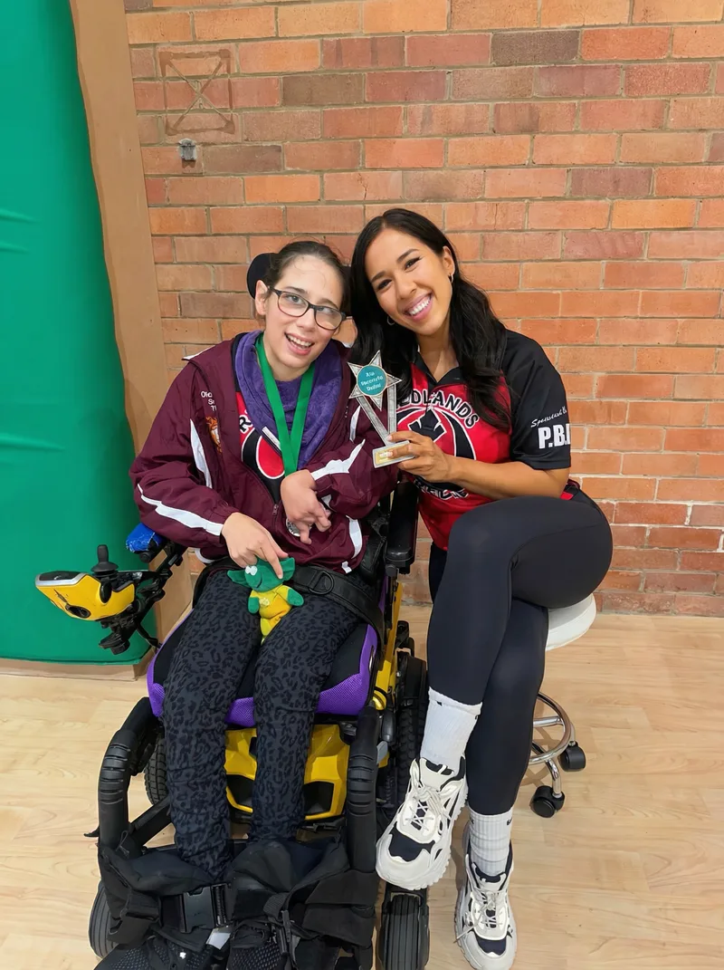 Danielle and Heidi holding the boccia trophy, both in Redlands Rebels shirts, grinning at the camera